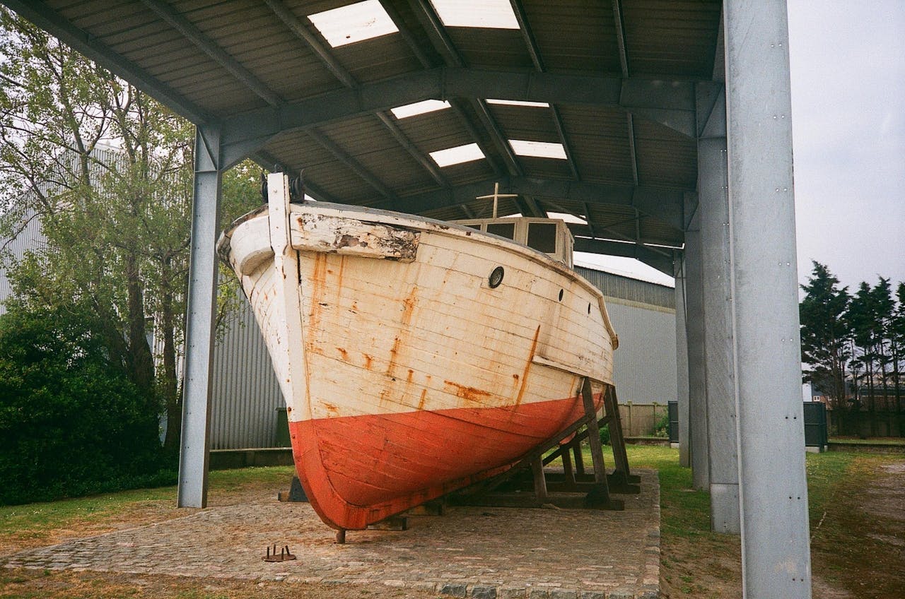 hero-img-01 A rusty, abandoned wooden boat under a metal shelter in Dunkirk, France.